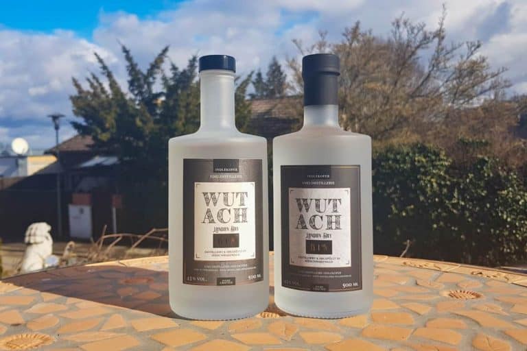 Zwei mattierte Flaschen „Wutach Japan Gin“ stehen auf einem sonnenbeschienenen Tisch im Freien, im Hintergrund sind Bäume und ein Haus vor einem teilweise bewölkten Himmel zu sehen.