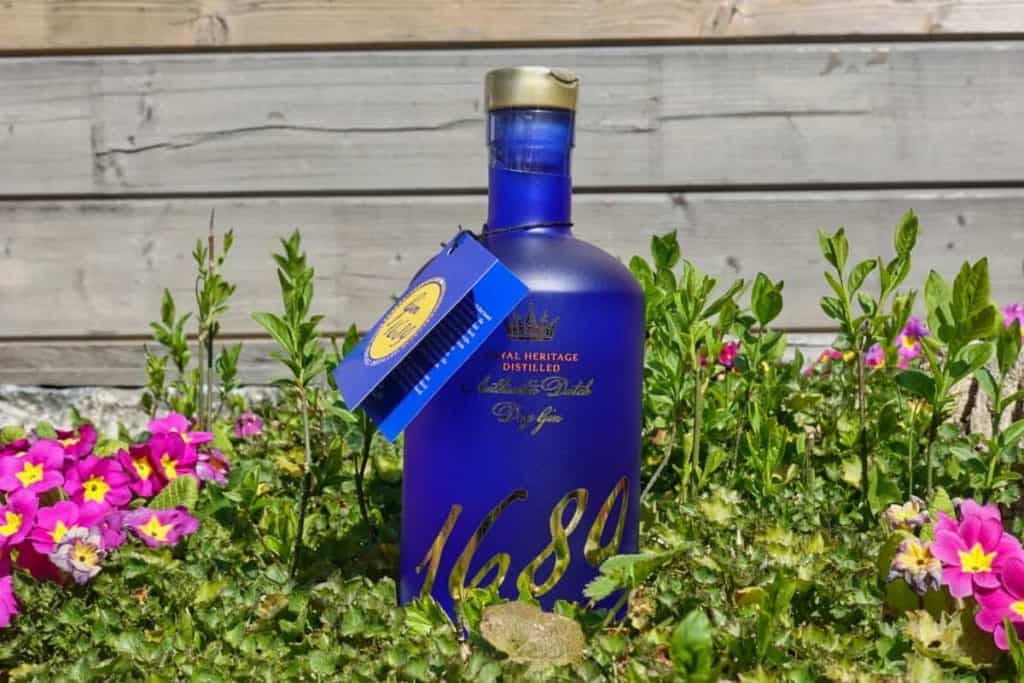 Zwischen grünen Pflanzen und rosa Blumen steht eine blaue Flasche mit der Aufschrift „1689“, im Hintergrund ist ein Holzzaun zu sehen. Am Flaschenhals hängt ein blau-gelbes Etikett.