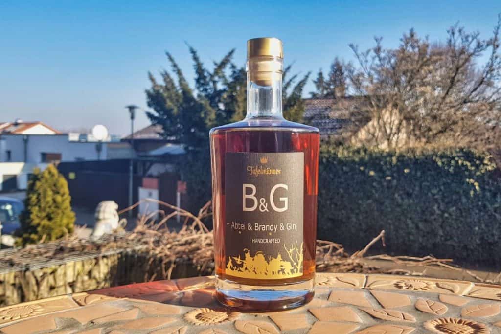 Auf einem Mosaiktisch im Freien steht eine Flasche „B&G Atelier Brandy & Gin“, im Hintergrund sind Häuser, Bäume und ein strahlend blauer Himmel zu sehen.