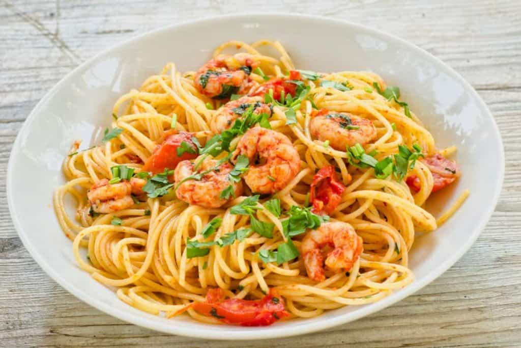 Eine weiße Schüssel, gefüllt mit Spaghetti, Garnelen und gewürfelten Tomaten, garniert mit gehackter Petersilie, steht auf einem rustikalen Holztisch.