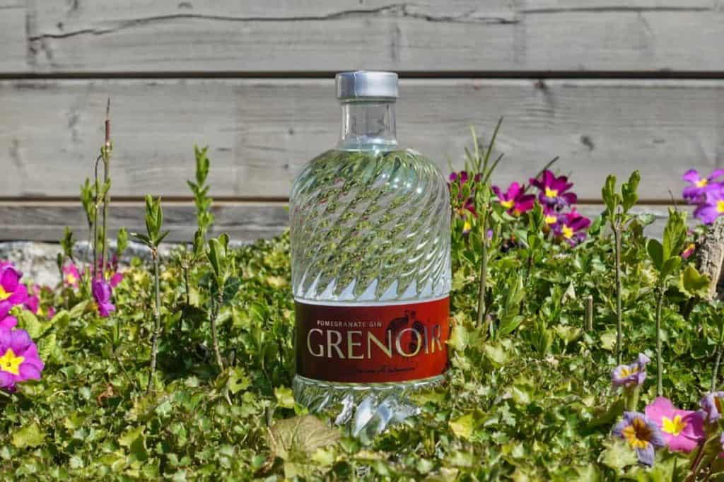Eine durchsichtige Flasche „Grenoir Pomegranate Gin“ steht inmitten von grünem Laub mit violetten und gelben Blüten vor einem Hintergrund aus Holz.