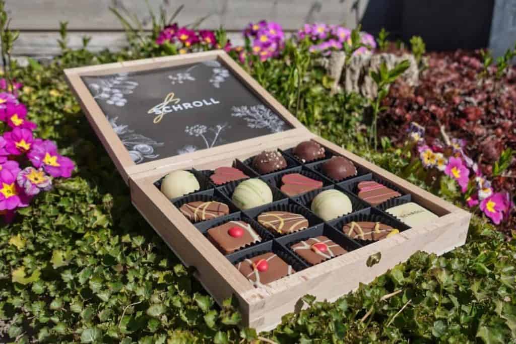 Eine Holzkiste mit verschiedenen Pralinen steht offen auf grünem Laub, umgeben von bunten rosa und gelben Blumen im Freien.