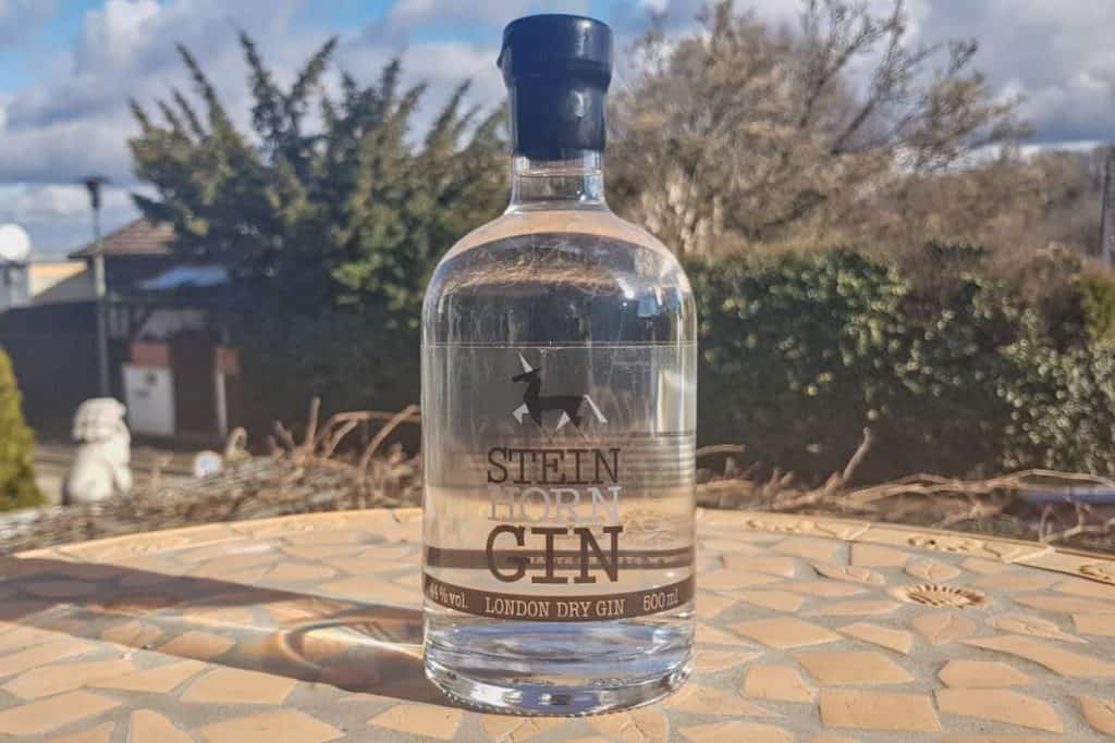 Auf einem Mosaiktisch im Freien steht eine durchsichtige Flasche „Stein Horn London Dry Gin“, im Hintergrund sind Bäume, ein Zaun und ein blauer Himmel mit Wolken zu sehen.