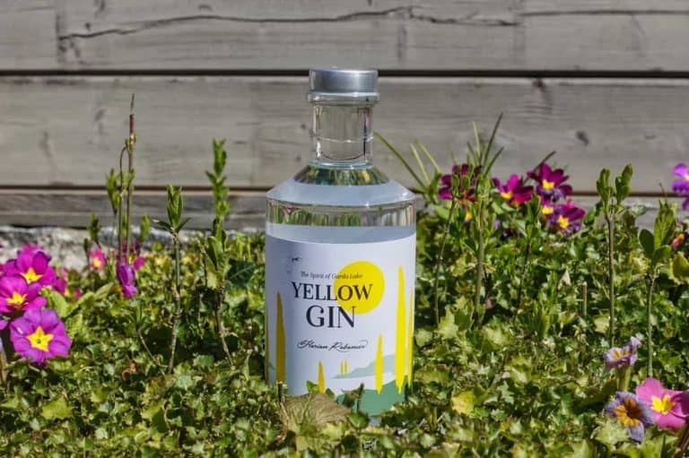 Auf grünem Gras steht eine Flasche „Yellow Gin“, umgeben von kleinen rosa und violetten Blumen, im Hintergrund ist ein Holzzaun zu sehen.