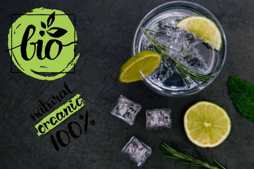 Ein Glas Sprudelwasser mit Eis, Limette und Rosmarin steht auf einer dunklen Unterlage, auf der Limettenscheiben, Minze und Eiswürfel liegen. Links steht in grüner und schwarzer Schrift „Bio 100 % natürlich und biologisch“.