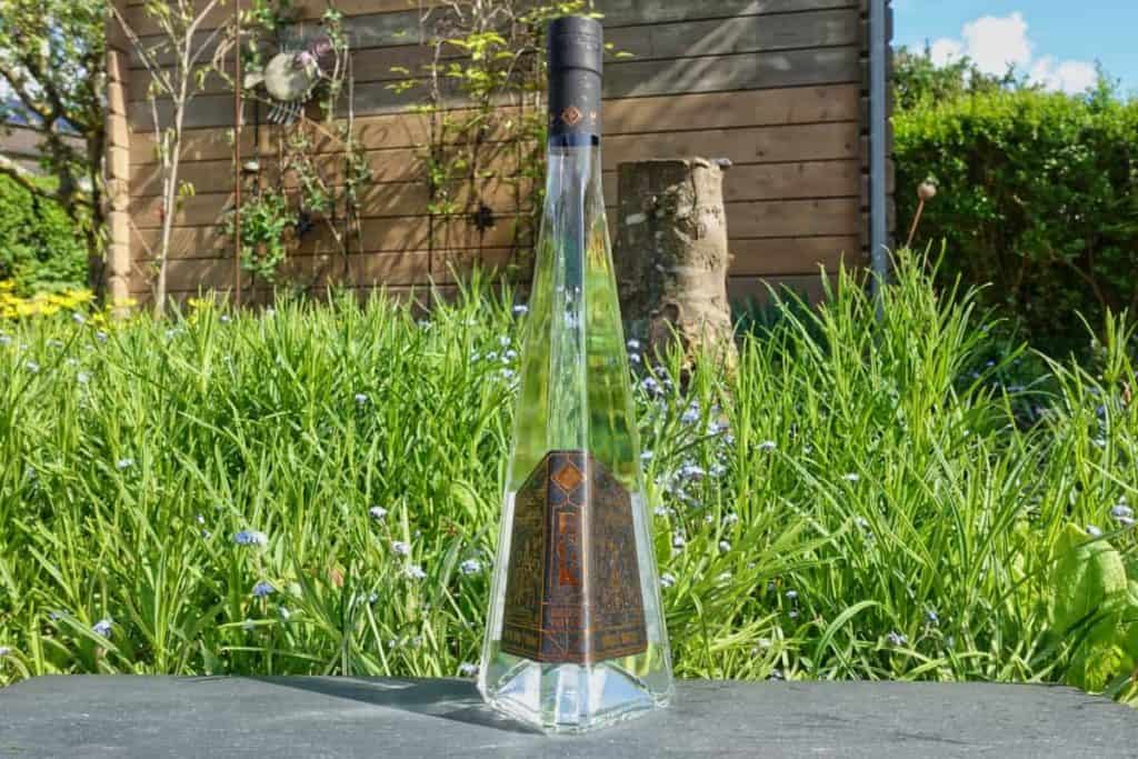 Eine hohe, einzigartig geformte, durchsichtige Flasche mit einem dunklen Verschluss steht im Freien auf einer Steinfläche, umgeben von grünem Gras und Pflanzen, im Hintergrund ist ein Holzzaun zu sehen.