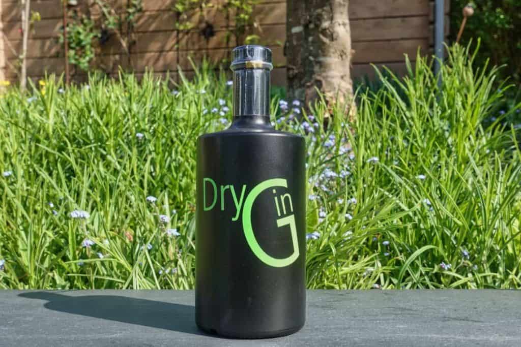 Eine schwarze Flasche mit der Aufschrift „Dry Gin“ in grüner Schrift steht im Freien auf einer grauen Fläche, im Hintergrund sind hohes grünes Gras und ein Holzzaun zu sehen.