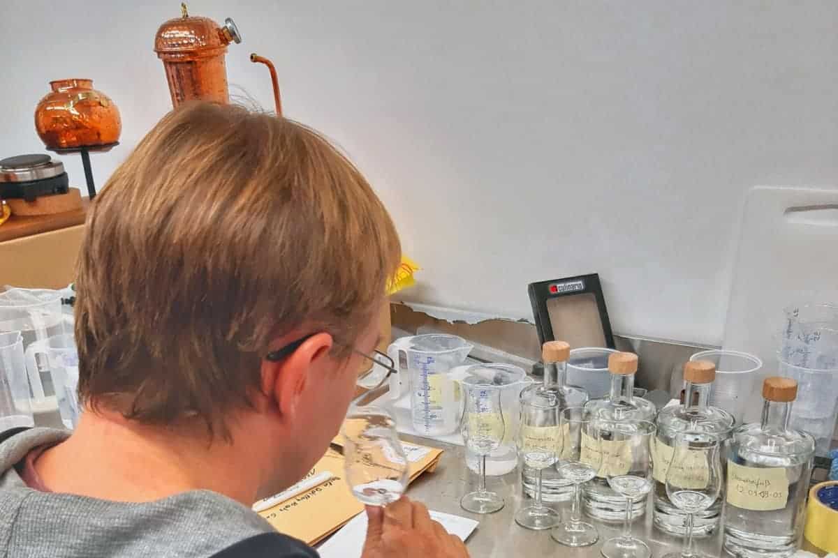 Eine Person mit hellbraunen Haaren riecht an einer klaren Flüssigkeit in einem Glas. Auf dem Tisch vor ihr stehen mehrere Glasflaschen mit Etiketten und Korken, Messbecher und Destilliergeräte.