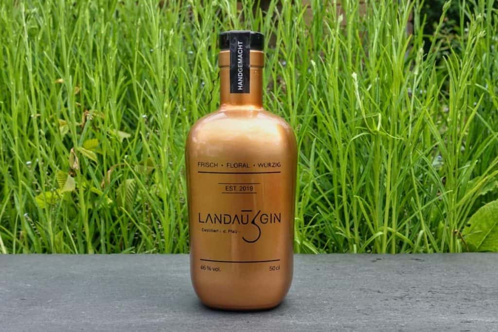 Auf einer grauen Fläche steht eine goldene Flasche Landau Gin mit schwarzer Beschriftung, im unscharfen Hintergrund sind hohes grünes Gras und Pflanzen zu sehen.