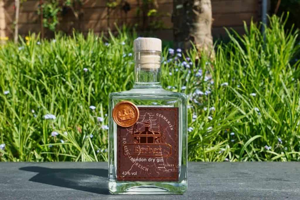 Eine Flasche Gin mit einem kupferfarbenen Etikett und einem Wachssiegel steht im Freien auf einer schwarzen Fläche, im Hintergrund sind grünes Gras und violette Blumen zu sehen.