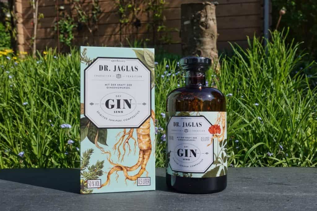 Eine Flasche Dr. Jaglas Gin Seng und ihre dekorative Schachtel stehen an einem sonnigen Tag im Freien auf einer Steinfläche, im Hintergrund sind grünes Gras, Blumen und ein Holzzaun zu sehen.