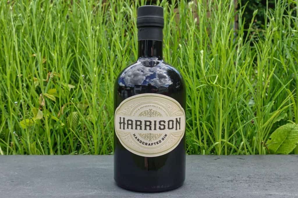 Auf einer grauen Fläche steht eine dunkle Glasflasche mit der Aufschrift „Harrison Handcrafted Gin“, im Hintergrund ist hohes grünes Gras zu sehen.