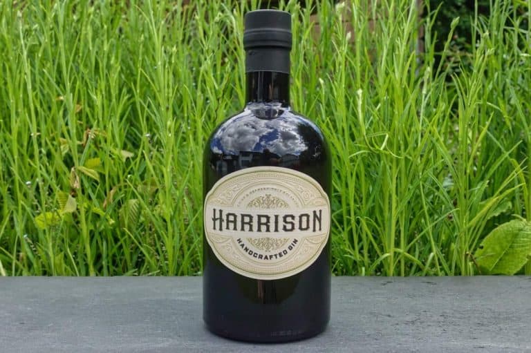Auf einer grauen Fläche steht eine dunkle Glasflasche mit der Aufschrift „Harrison Handcrafted Gin“, im Hintergrund ist hohes grünes Gras zu sehen.