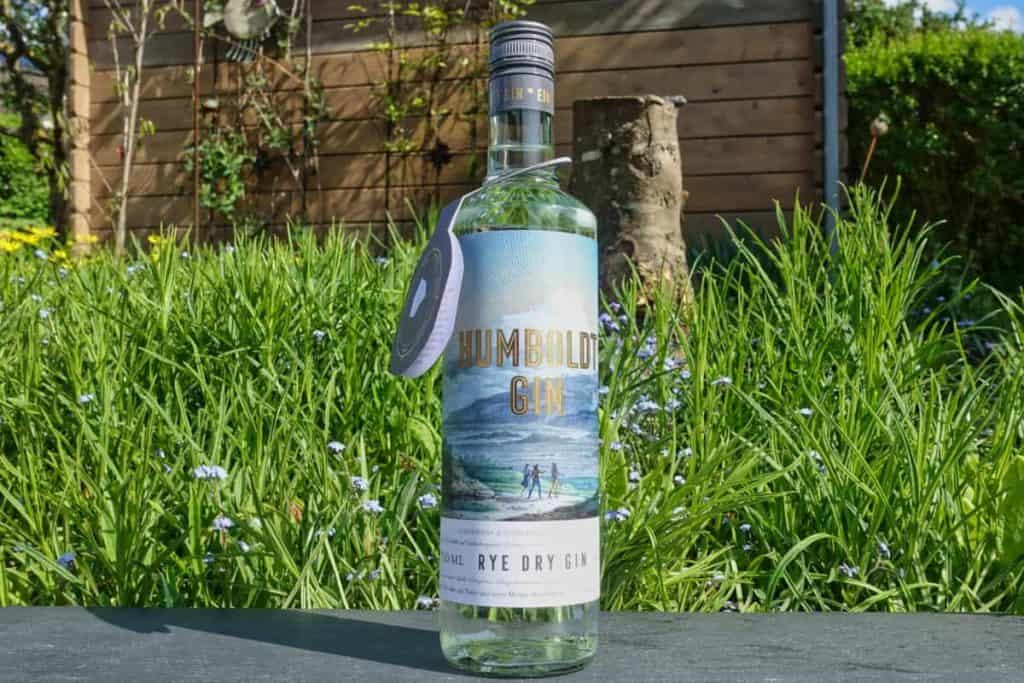 Eine Flasche Humboldt Gin steht im Freien auf einer Fläche, im Hintergrund sind an einem sonnigen Tag hohes grünes Gras, kleine blaue Blumen und ein Holzzaun zu sehen.