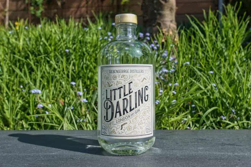 Eine durchsichtige Glasflasche mit der Aufschrift „Little Darling London Gin“ steht im Freien auf einer dunklen Fläche, im Hintergrund sind grünes Gras und kleine blaue Blumen zu sehen. Die Flasche ist teilweise gefüllt und hat einen hellen Verschluss.