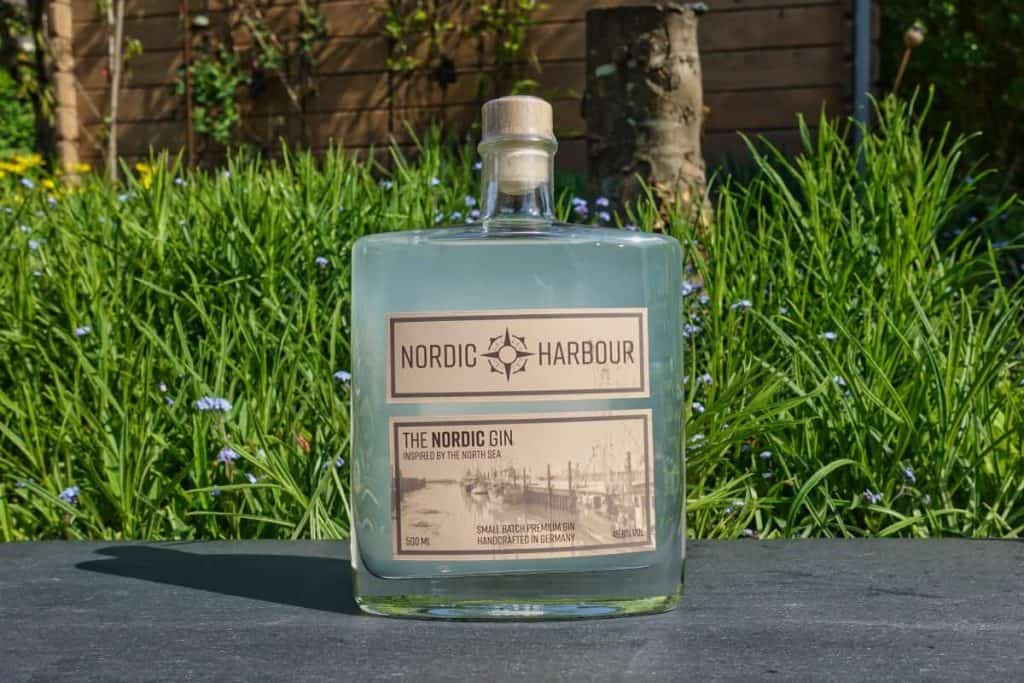 Eine Flasche „Nordic Harbour“-Gin steht im Freien auf einer dunklen Unterlage, im Hintergrund sind grüne Pflanzen und violette Blumen zu sehen. Das Etikett ist beige und mit schwarzer Schrift und Grafiken versehen.