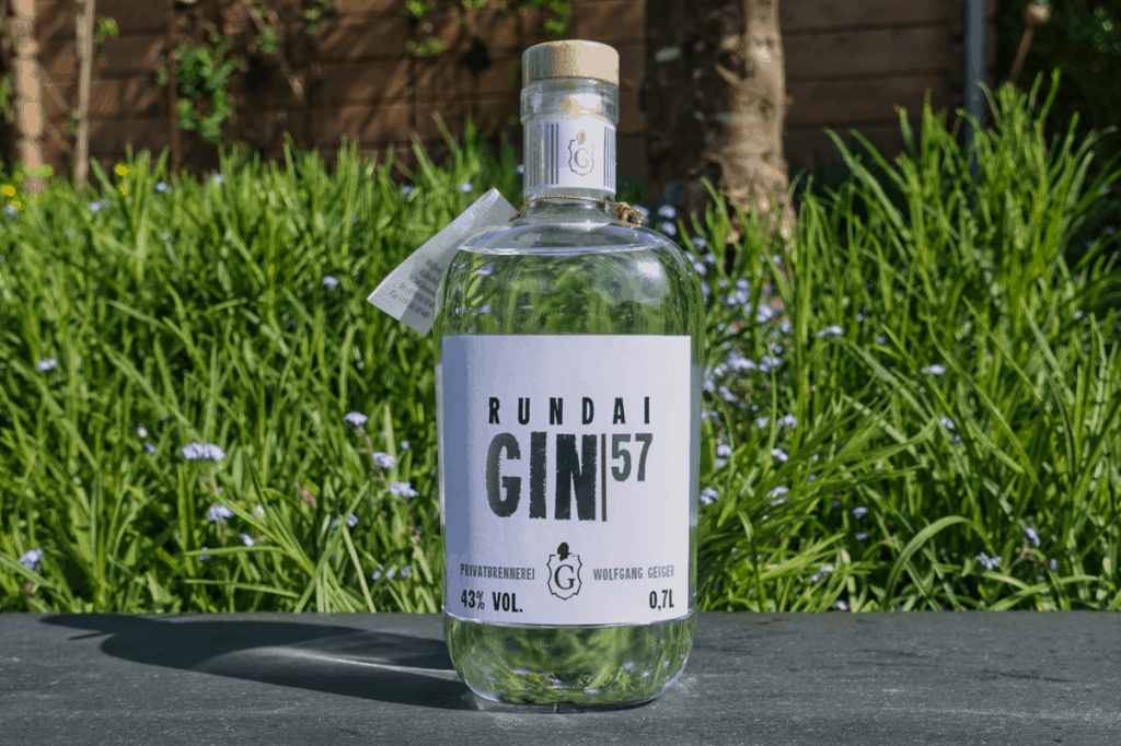 Eine durchsichtige Flasche „Rundai Gin 57“ steht im Freien auf einer dunklen Fläche, im Hintergrund sind grünes Gras und kleine violette Blüten zu sehen.