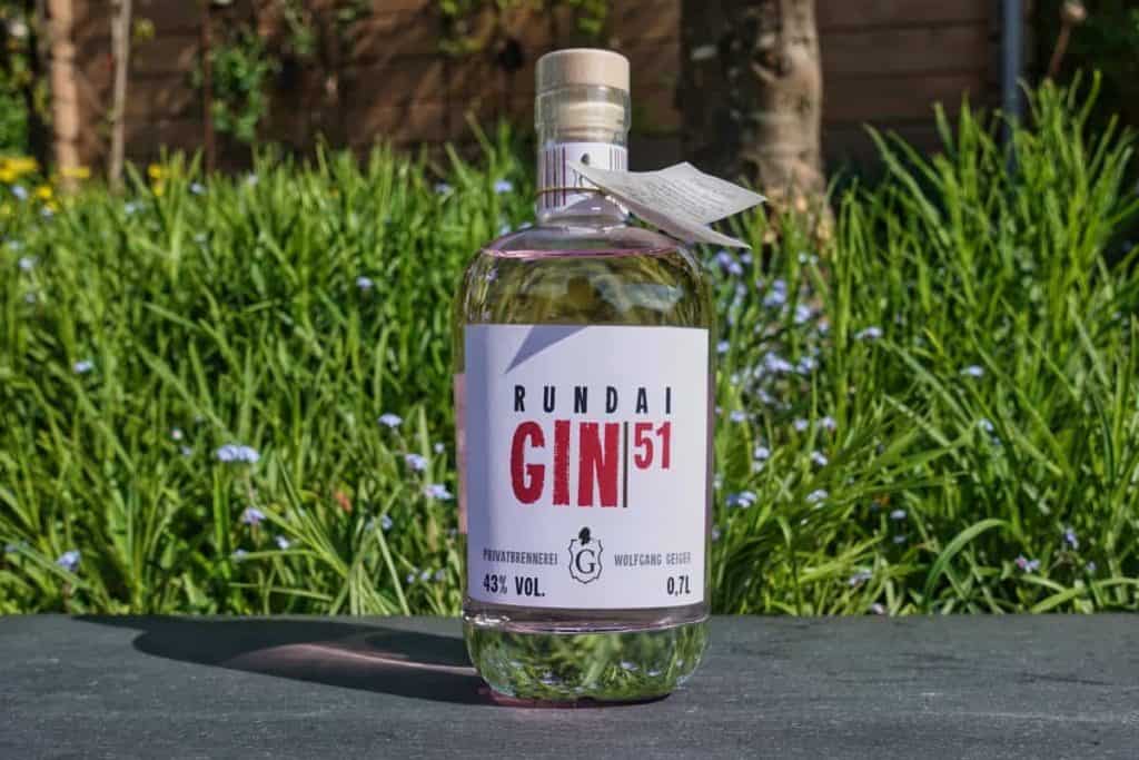 Eine Flasche Rundai Gin 51 steht im Freien auf einer dunklen Unterlage, im Hintergrund sind grünes Gras und violette Blumen zu sehen. Auf dem Etikett sind der Markenname, der Alkoholgehalt und das Volumen angegeben.