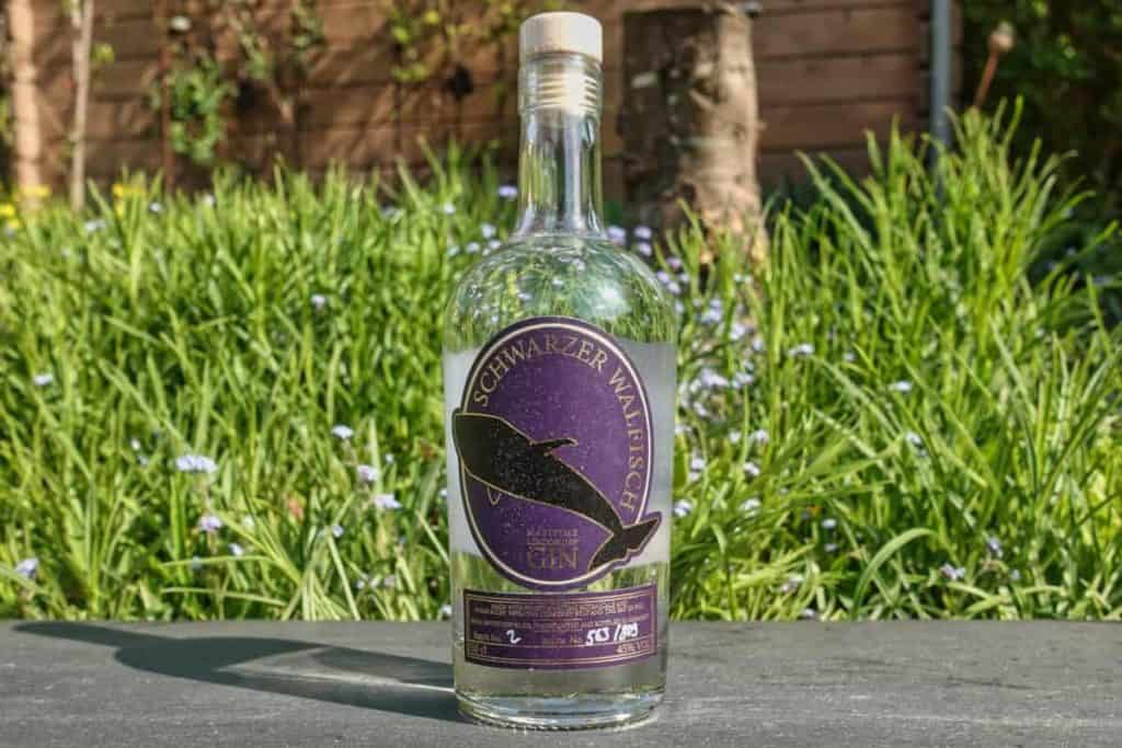 Eine durchsichtige Flasche „Schwarzer Walfisch“-Gin mit einem dunklen Etikett, auf dem ein Wal abgebildet ist, im Freien auf einer Steinfläche platziert, im Hintergrund grünes Gras und ein Holzzaun.