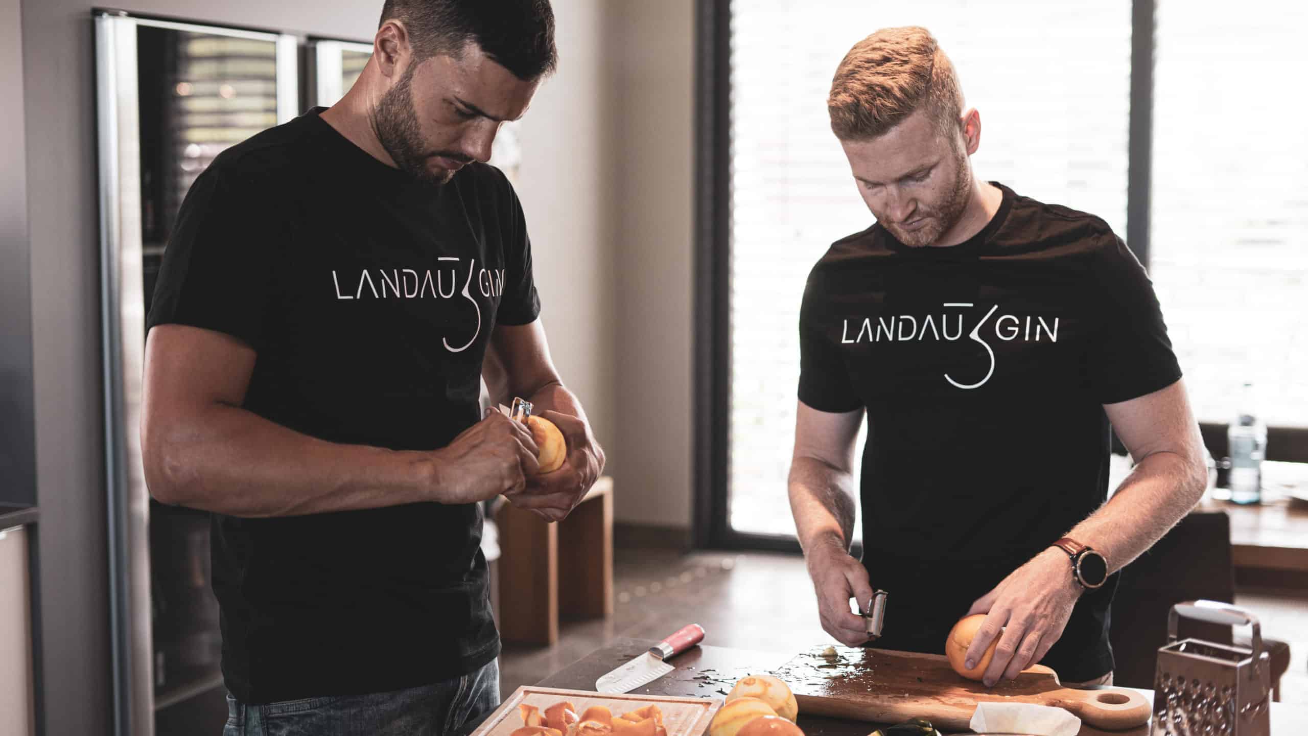 Zwei Männer, die identische Landau-Gin-Shirts tragen, schälen und schneiden in einer modernen Küche Orangen und sind ganz auf ihre Aufgabe konzentriert.