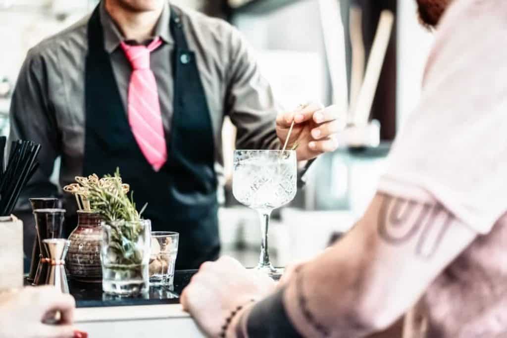 Ein Barkeeper in einer schwarzen Weste und einer rosa Krawatte rührt einen Cocktail in einem großen Glas um, während ein Gast mit tätowierten Armen von der anderen Seite der Bar aus zusieht. Auf der Theke liegen Kräutergarnituren und Barutensilien.