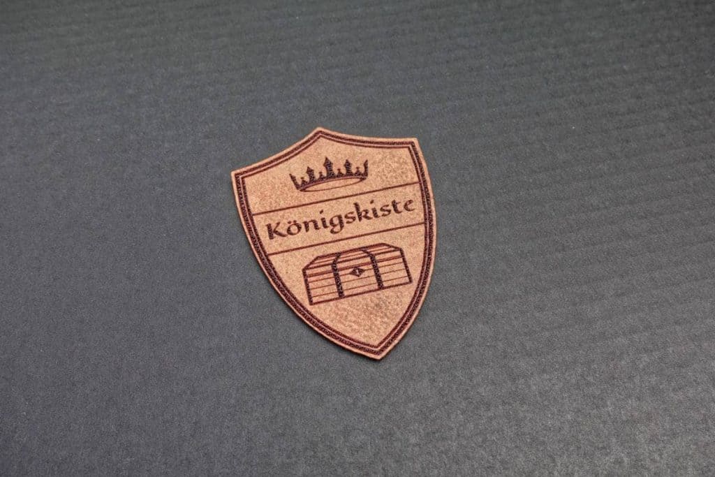 Ein brauner, schildförmiger Aufdruck mit einer Krone, dem Wort „Königskiste“ und der Abbildung einer Truhe auf einem dunklen, strukturierten Hintergrund.