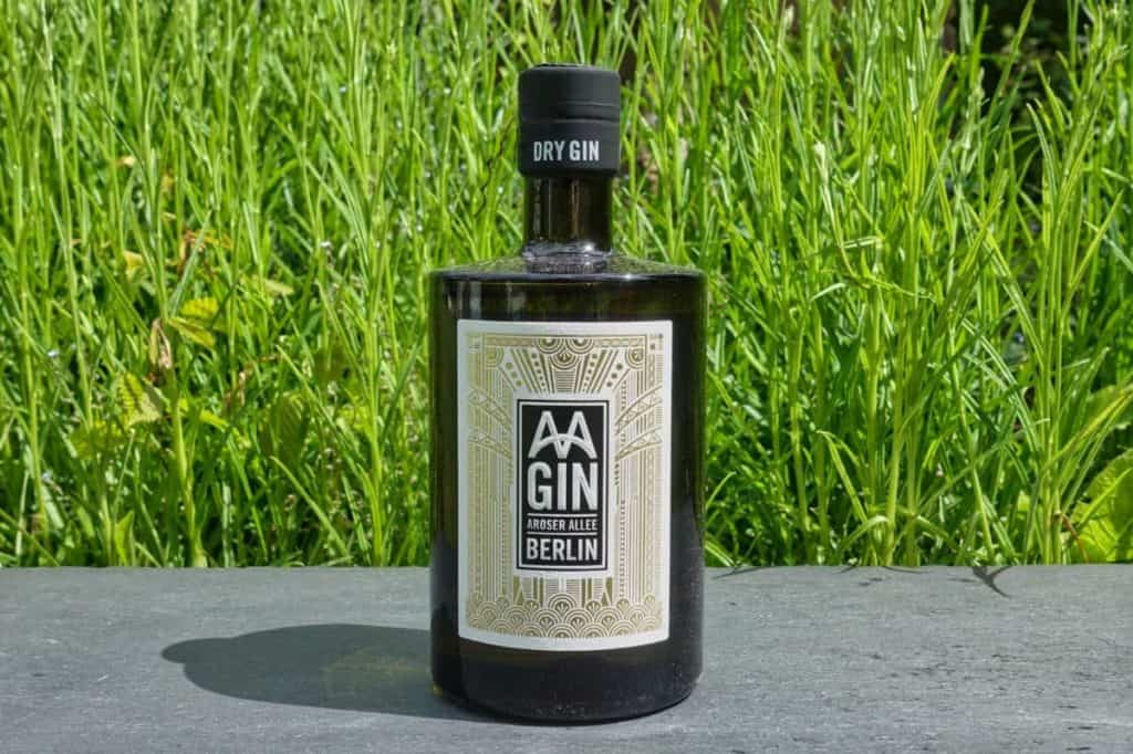 Eine schwarze Flasche AA Gin mit der Aufschrift „Dry Gin BERLIN“ steht auf einer Steinfläche, im Hintergrund ist hohes grünes Gras zu sehen.
