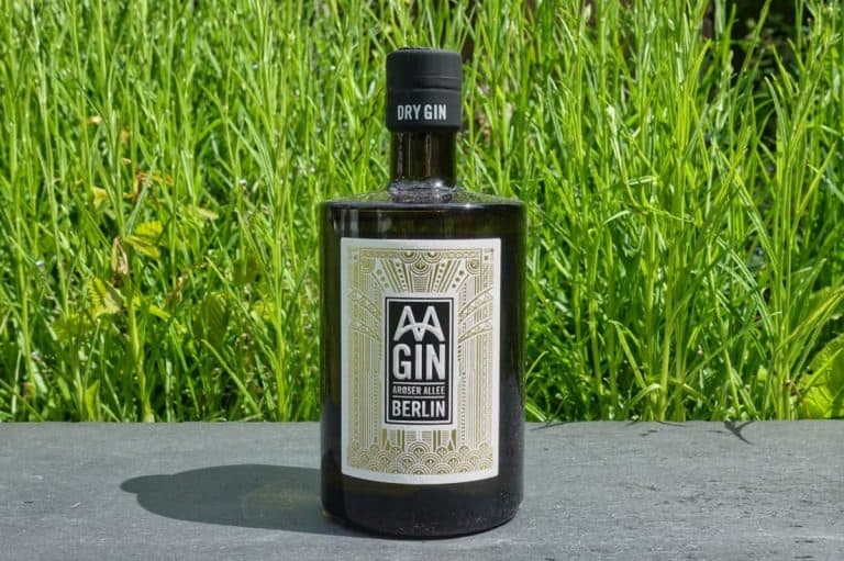 Eine schwarze Flasche AA Gin mit der Aufschrift „Dry Gin BERLIN“ steht auf einer Steinfläche, im Hintergrund ist hohes grünes Gras zu sehen.