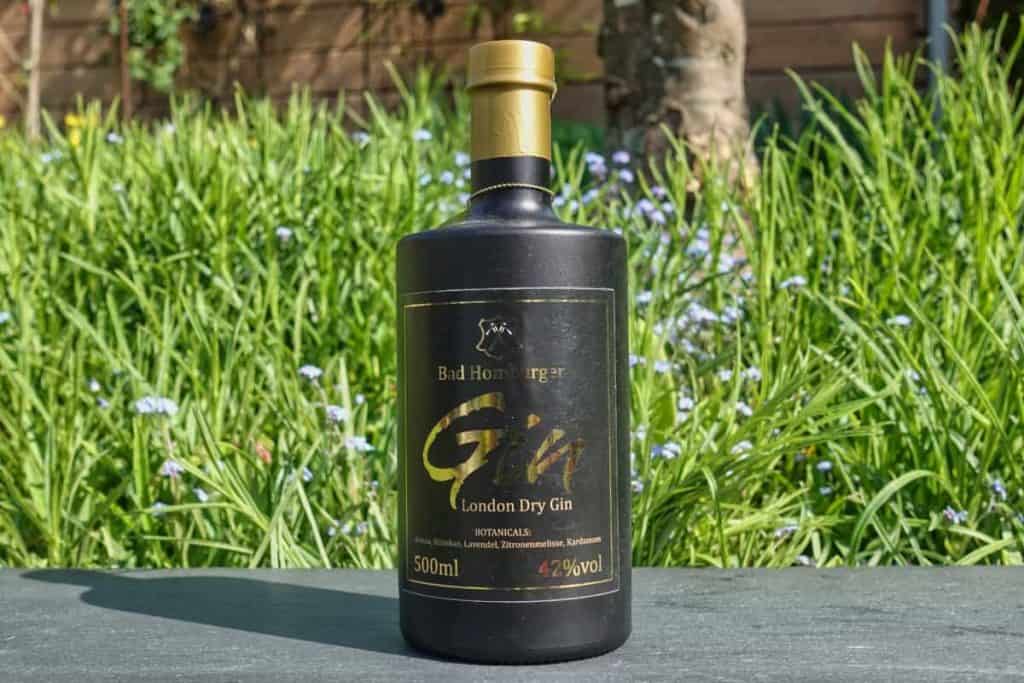 Eine schwarze Flasche „Bad Homburger London Dry Gin“ steht auf einem Tisch im Freien, im Hintergrund sind an einem sonnigen Tag grünes Gras und violette Blumen zu sehen.
