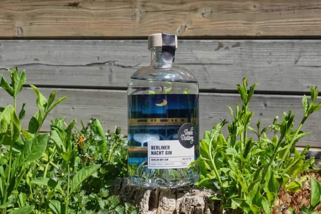 Auf einem Baumstumpf inmitten grüner Pflanzen steht eine Flasche „Berliner Nacht Gin“, im Hintergrund ist ein Holzzaun zu sehen.