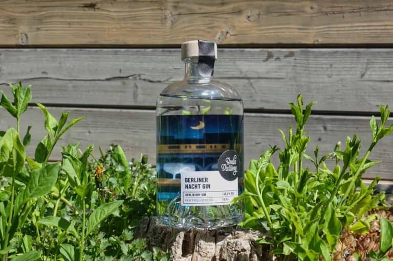 Auf einem Baumstumpf inmitten grüner Pflanzen steht eine Flasche „Berliner Nacht Gin“, im Hintergrund ist ein Holzzaun zu sehen.