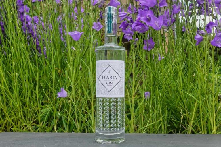 Eine Flasche DAria Renosterbos Gin steht im Freien auf einer dunklen Fläche, im Hintergrund sind grünes Gras und leuchtend violette Blumen zu sehen.