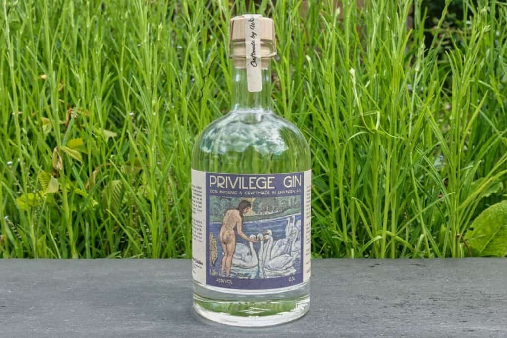 Eine Flasche „Privilege Gin“ mit Korkverschluss steht auf einer dunklen Fläche vor hohem grünem Gras. Auf dem Etikett ist eine Person abgebildet, die neben Schwänen steht.