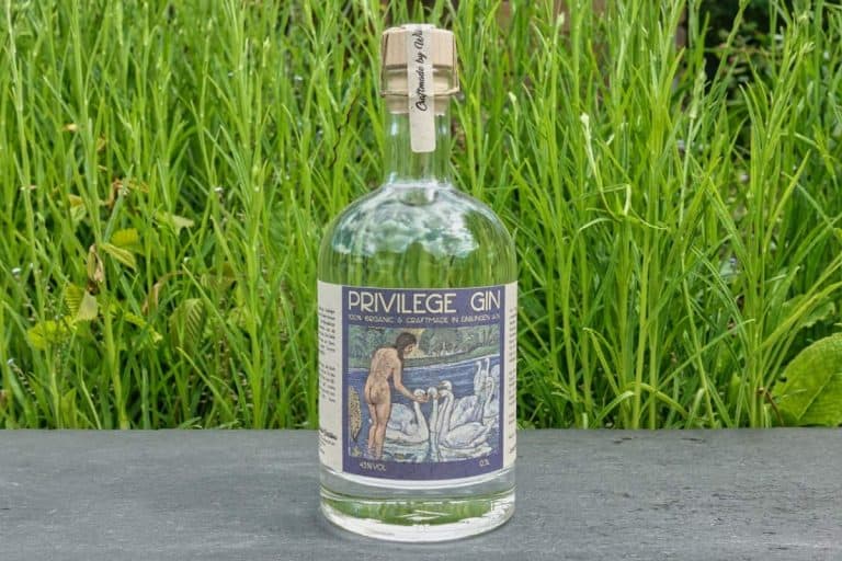 Eine Flasche „Privilege Gin“ mit Korkverschluss steht auf einer dunklen Fläche vor hohem grünem Gras. Auf dem Etikett ist eine Person abgebildet, die neben Schwänen steht.