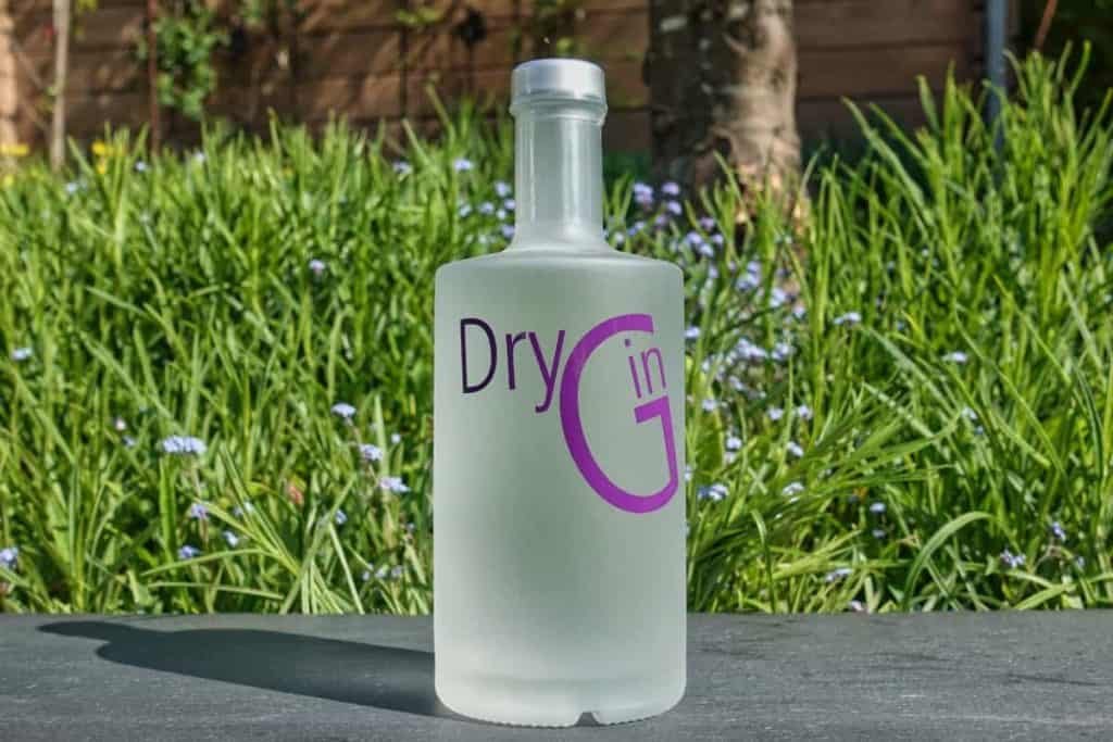 Eine mattierte Flasche mit der Aufschrift „Dry Gin“ in violetten Buchstaben steht im Freien auf einer grauen Fläche, im Hintergrund sind grünes Gras und violette Blumen zu sehen.