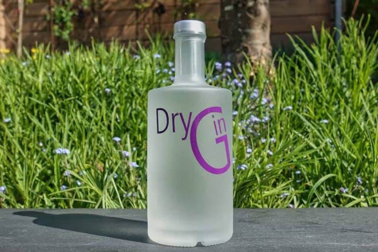 Eine mattierte Flasche mit der Aufschrift „Dry Gin“ in violetten Buchstaben steht im Freien auf einer grauen Fläche, im Hintergrund sind grünes Gras und violette Blumen zu sehen.
