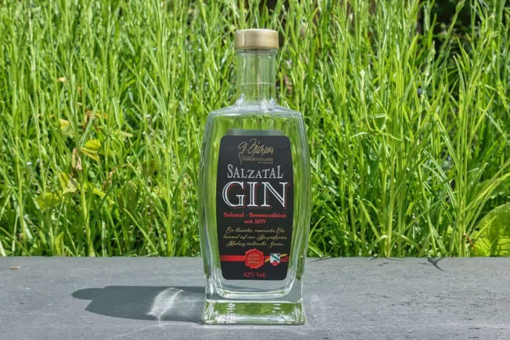 Eine durchsichtige Glasflasche mit Salztal Gin und schwarzem Etikett steht auf einer grauen Fläche vor dem Hintergrund von hohem, grünem Gras an einem sonnigen Tag.