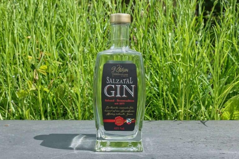Eine durchsichtige Glasflasche mit Salztal Gin und schwarzem Etikett steht auf einer grauen Fläche vor dem Hintergrund von hohem, grünem Gras an einem sonnigen Tag.