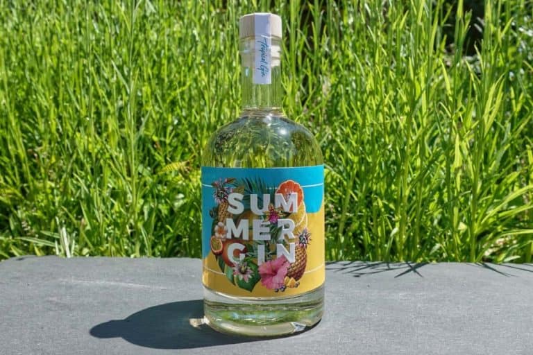 Eine Flasche „Summer Gin“ mit einem farbenfrohen Etikett im tropischen Stil steht auf einer dunklen Unterlage, wobei das üppig grüne Gras im Hintergrund den lebhaften Charakter des „Summer Gin“ perfekt unterstreicht.