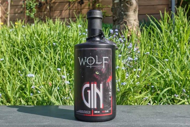 Eine schwarze Flasche Wolf Gin steht im Freien auf einer Steinfläche, im Hintergrund sind hohes grünes Gras und ein Holzzaun zu sehen. Das Etikett zeigt das Gesicht eines Wolfes sowie auffällige weiße und rote Schriftzüge und fängt mit seinem markanten Design den Charakter von Wolf Gin perfekt ein.
