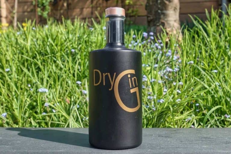 Eine mattschwarze Flasche mit der Aufschrift „Gin Gold“ in schimmernden Buchstaben steht im Freien auf einer Schieferfläche, im Hintergrund sind hohes grünes Gras und kleine violette Blumen zu sehen.