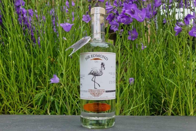 Eine Flasche Sir Edmond Gin mit einer Vogelillustration auf dem Etikett steht auf einer Fläche vor dem Hintergrund von hohem grünem Gras und blühenden violetten Blumen und fängt damit perfekt die natürliche Eleganz ein, die hinter jedem Glas Sir Edmond Gin steckt.
