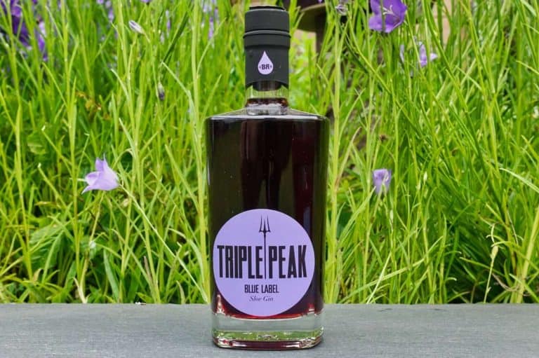 Eine Flasche „Triple Peak Blue Label Sloe Gin“, deren markantes Gin-Etikett nach vorne zeigt, steht im Freien auf einer Fläche, im Hintergrund sind hohes grünes Gras und violette Blumen zu sehen.