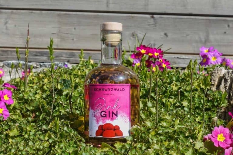 Auf grünem Gras steht eine Flasche Pink Gin, umgeben von kleinen rosa und gelben Blumen, im Hintergrund ist ein Holzzaun zu sehen.