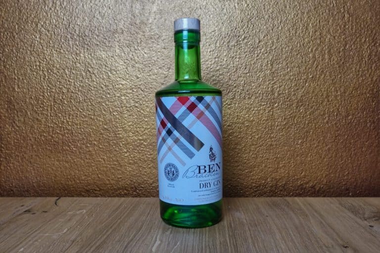 Eine grüne Glasflasche „3Bens Bracovniça Dry Gin“ mit einem gemusterten weißen Etikett steht auf einer Holzfläche vor einem strukturierten braunen Hintergrund, was an den unverwechselbaren Stil des „Ben Bracken Dry Gin“ (Lidl) erinnert.
