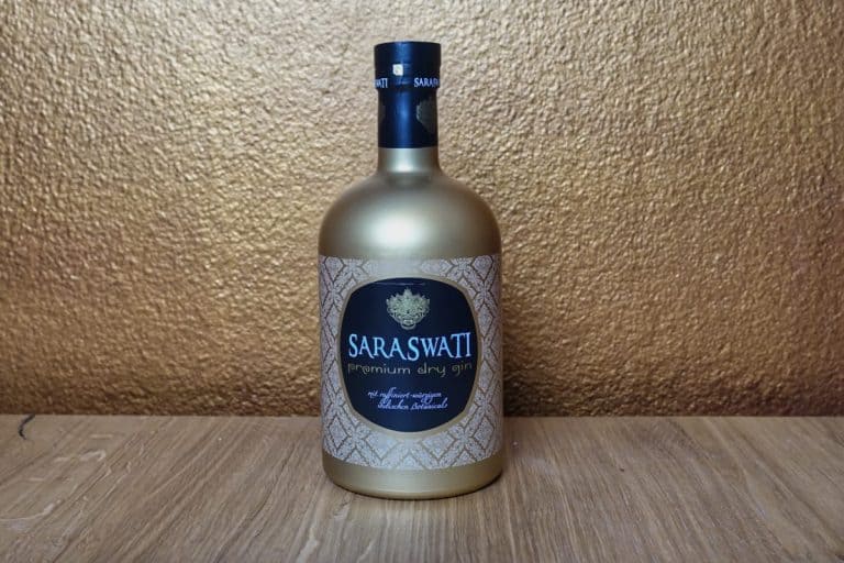 Eine goldene Flasche „Saraswatti Premium Dry Gin“ steht auf einer Holzfläche, im Hintergrund ist eine strukturierte goldene Wand zu sehen.
