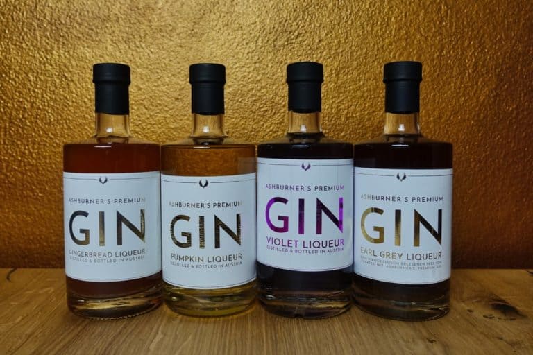 Vier Flaschen „Ashburners Premium Gin Liqueur“ – in den Geschmacksrichtungen Lebkuchen, Kürbis, Veilchen und Earl Grey – stehen auf einer Holzunterlage vor einem goldenen Hintergrund in einer Reihe.