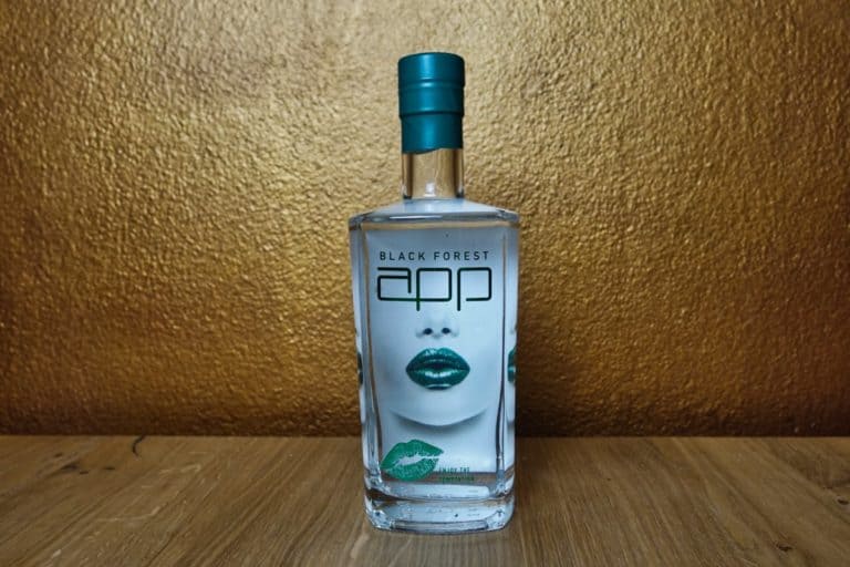 Eine klare Flasche mit Schwarzwälder Likör, versehen mit einem blaugrünen Verschluss, auf deren Etikett ein Bild von Lippen mit grünem Lippenstift zu sehen ist. Inspiriert von der Faszination des Black Gin steht die Flasche auf einer Holzunterlage vor einem goldfarbenen, strukturierten Hintergrund.