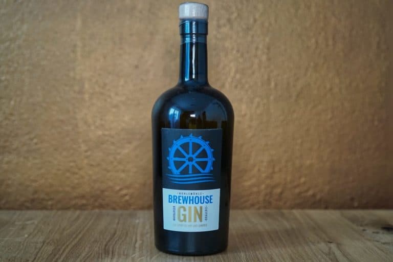 Eine dunkle Glasflasche mit blauem Verschluss und einem Etikett mit der Aufschrift „Warburger Brewhouse Gin“ steht auf einer Holzunterlage vor einem strukturierten, braunen Hintergrund.