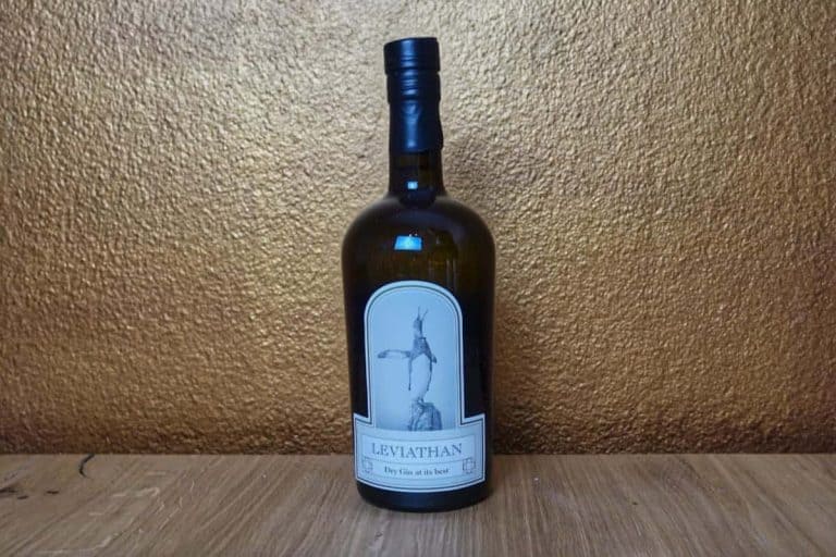Eine Flasche Leviathan London Dry Gin steht auf einer Holzfläche vor einem strukturierten goldenen Hintergrund. Auf dem Etikett ist eine Illustration eines Wesens zu sehen, das aus dem Wasser auftaucht.